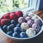 Fresh berries including strawberries, blueberries, and three types of energy bites with oat and berry toppings in a white bowl on a wooden table by a window.