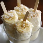 Creamy banana ice cream with coconut flakes garnished with wooden popsicle sticks, served in a glass bowl. Perfect for summer treats and homemade dessert recipes.
