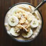 Banana and peanut butter smoothie topped with banana slices and shredded coconut, served in a clear glass bowl with a spoon, on a wooden surface.