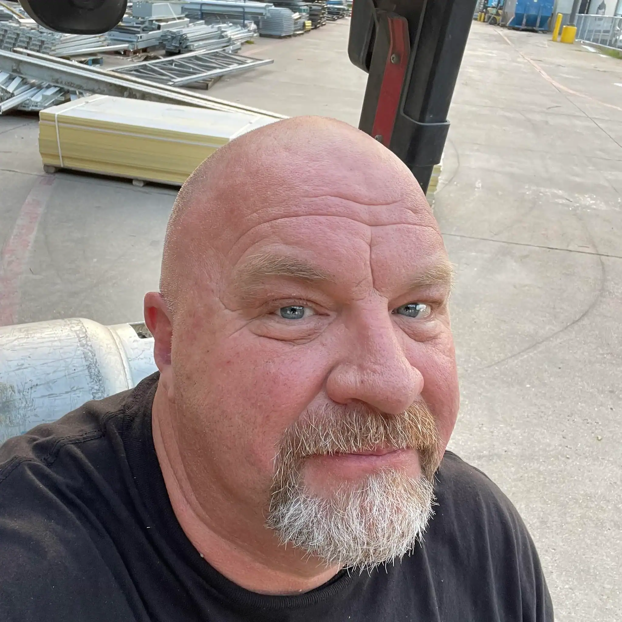 Shaved bald man with a beard and blue eyes taking a selfie outdoors at a warehouse or industrial yard with metal shelving and construction materials in the background.