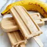 Creamy banana flavored dog ice cream bars with vanilla flavor and wooden sticks, served with a fresh banana on marble background. Perfect homemade frozen treat for dogs on hot days.