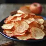 Crispy homemade apple chips on a black plate, seasoned with cinnamon, served as a healthy snack with fresh apples in the background. Perfect for healthy eating or dessert.