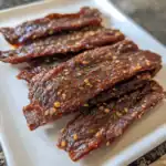Thinly sliced beef jerky with sesame seeds on a white platter, perfect for snacks or outdoor trips. Handmade Waggy Chef beef jerky offers rich flavor, tender texture, and high-quality ingredients.